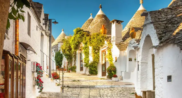 Un vicolo incantevole tra i trulli, perfetti borghi da visitare in Puglia, con fiori rossi e rampicanti verdi.